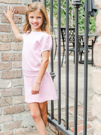 Young girl in a pink dress standing against a brick wall with outdoor furniture in the background.