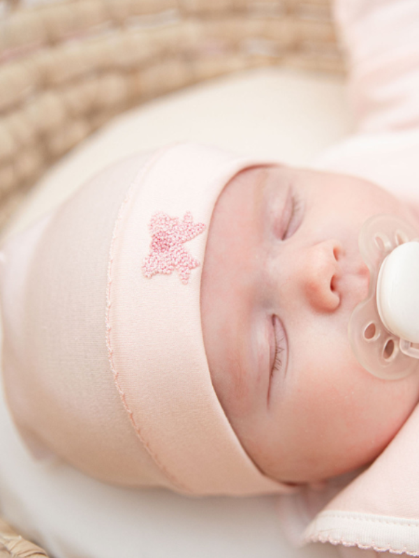 A sleeping infant wearing a light pink Pima cotton baby hat with a hand-embroidered bow and picot trim.