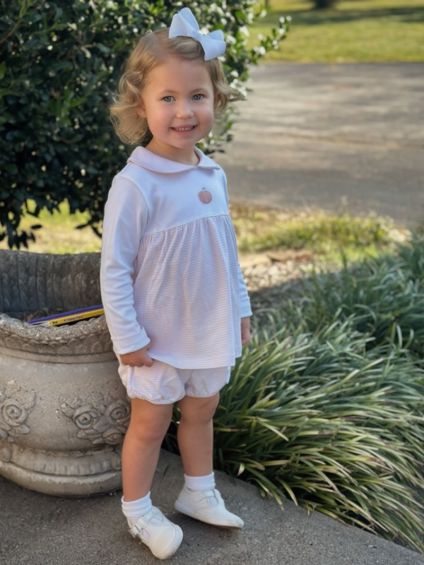 Young girl in a white and pink stripe bloomer set with a pumpkin embroidery on top standing outdoors.