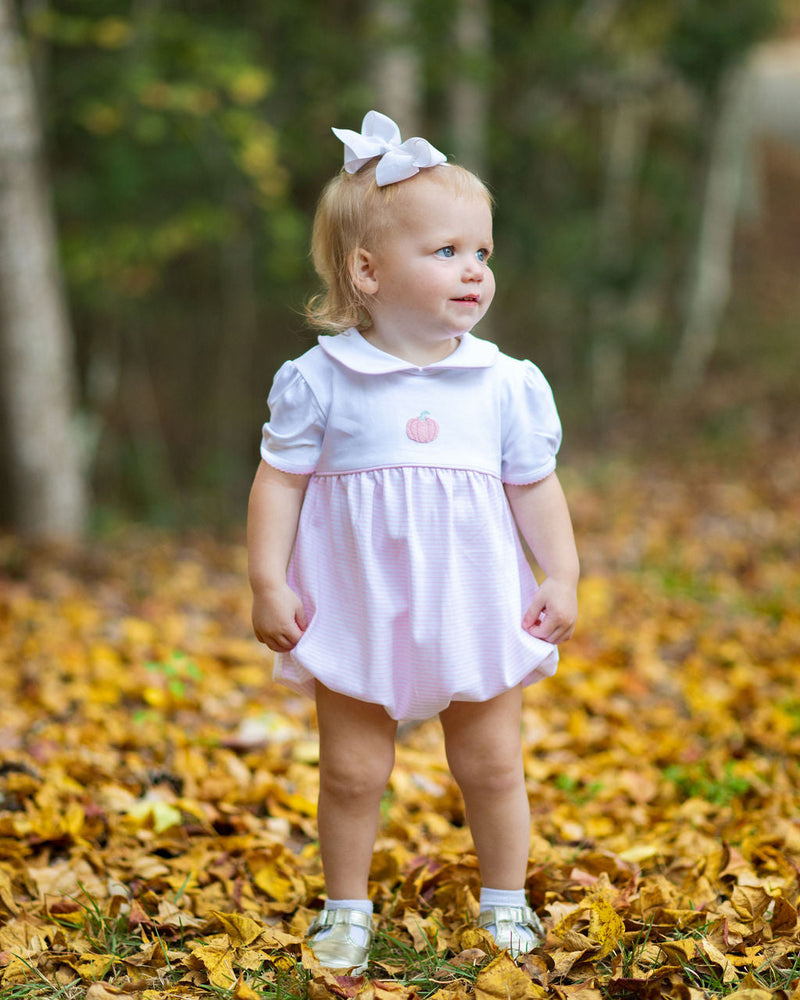 Young girl in a white and pink stripe baby bubble with a pink pumpkin standing on autumn leaves.