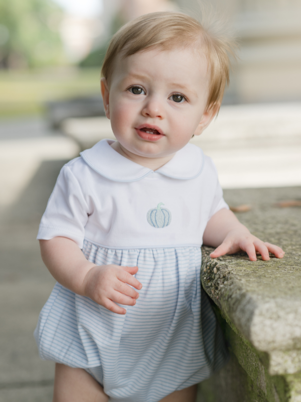 Baby in a white and blue stripe Pima cotton bubble with hand embroidered pumpkin standing on a stone surface.