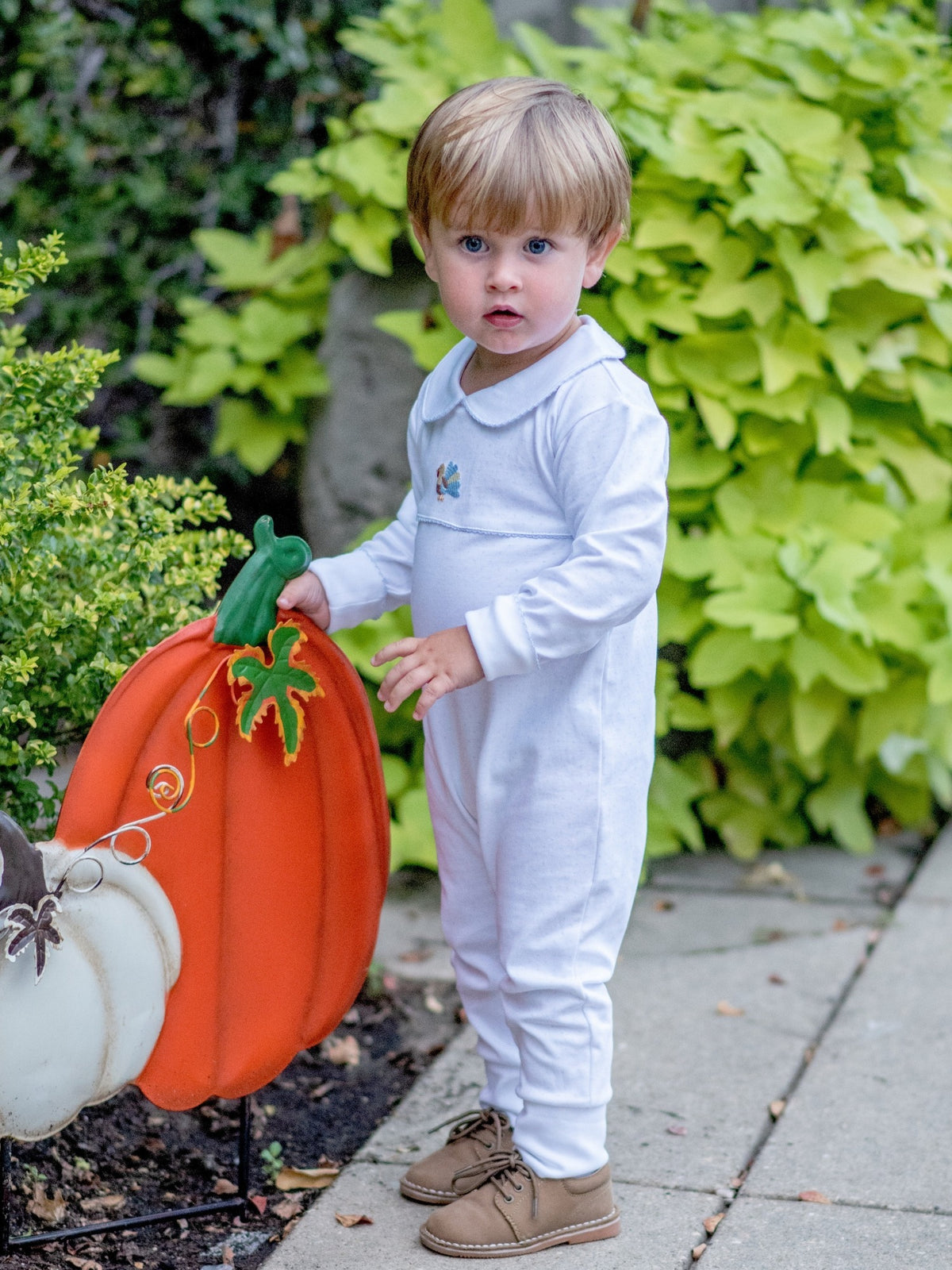 Child in a white playsuitoutfit holding a pumpkin-shaped sign outdoors.