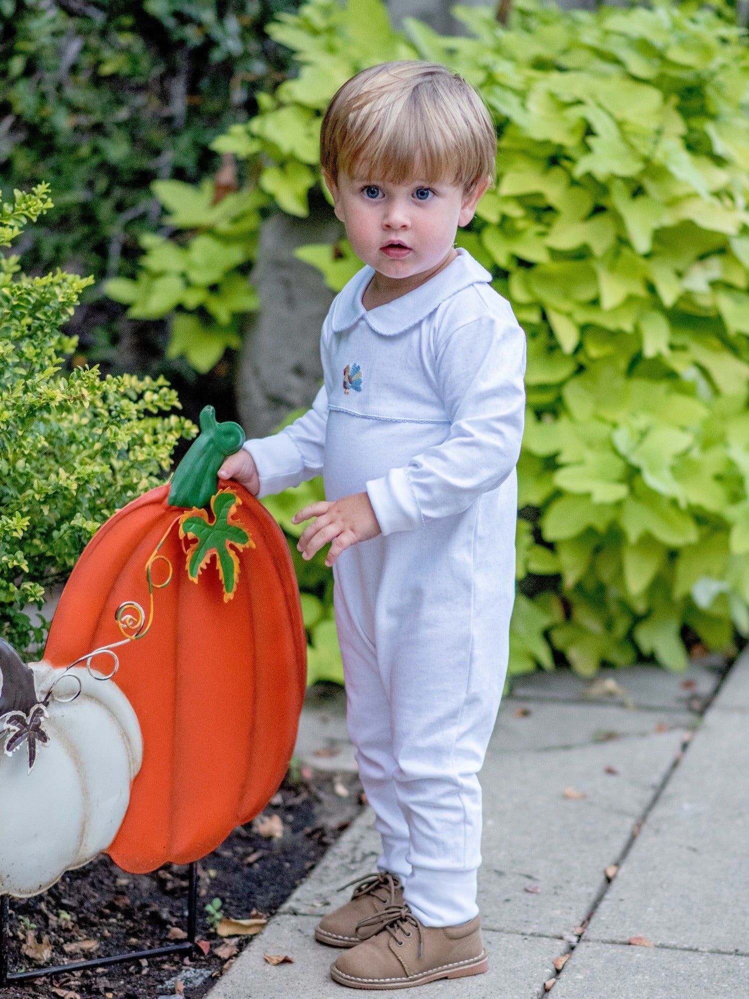 Child in a white playsuitoutfit holding a pumpkin-shaped sign outdoors.