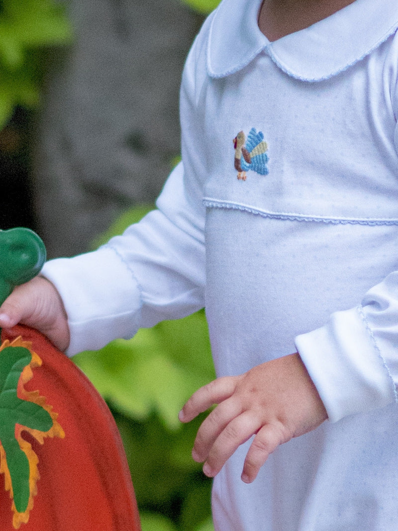 Child wearing a white blue bitty dot playsuit/romper with a embroidered turkey design, holding a pumpkin.
