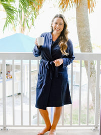 Woman in a navy robe holding a white mug on a balcony with palm trees in the background