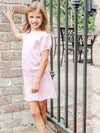 Young girl in a pink dress standing against a brick wall with outdoor furniture in the background.