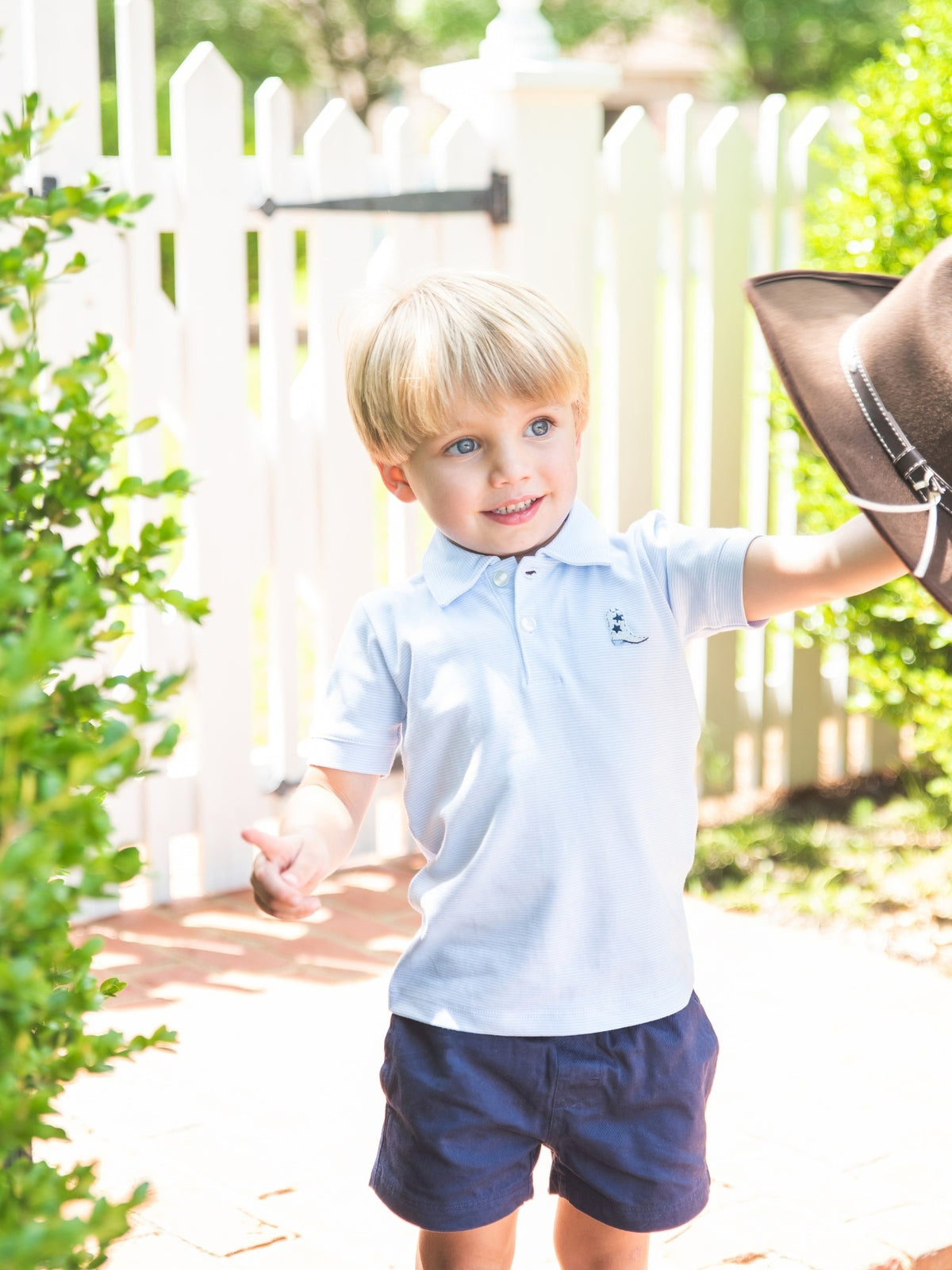A young blond boy wearing a light blue polo shirt and navy shorts stands outdoors in front of a white picket fence, holding a brown cowboy hat.