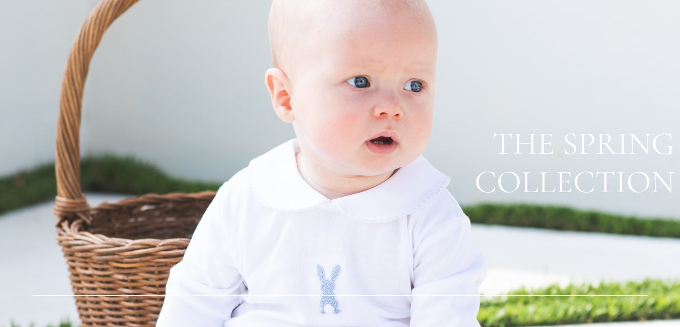 Baby in a white outfit with a blue bunny design, sitting next to a wicker basket with a spring theme.